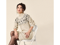 Woman in a beige patterned knit dress and black tights, sitting on a chair.