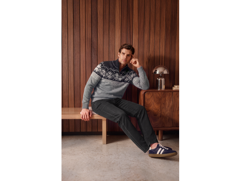 Man in a grey and black patterned half-zip sweater and dark jeans, sitting on a bench.