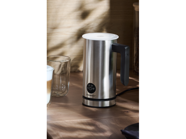 Silver milk frother on a wooden table, with a latte and an empty glass next to it.