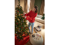 Woman decorates Christmas tree with lights and ornaments, gifts and decorations on pouf.