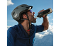 Man in helmet and sportswear drinking water from a bottle, with mountains in the background.