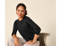 Woman in a black long-sleeved top and light trousers, sitting on a chair.