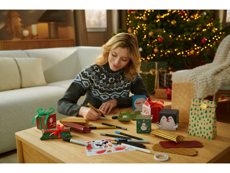 Woman preparing Christmas gift packaging, with a Christmas tree in the background.