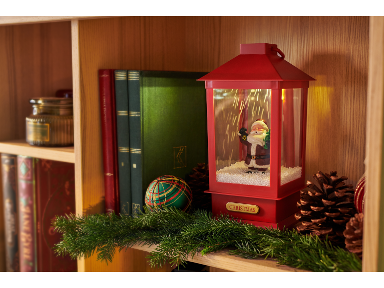 Christmas decoration with red lantern featuring Santa, books, and garland on a wooden shelf.