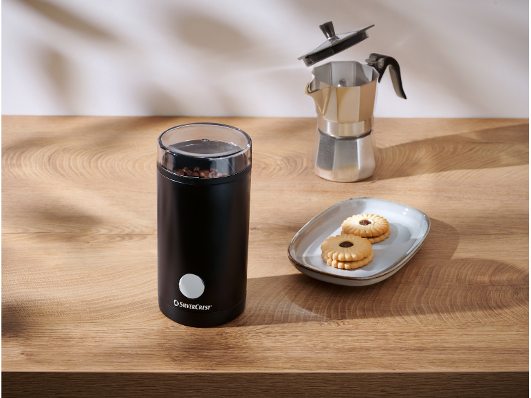 Black coffee grinder with beans, moka pot, and biscuits on a wooden table.