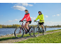Two smiling people on bicycles on a path by the water on a sunny day.