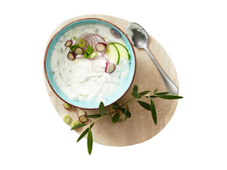 Tzatziki with cucumber, red onion, and spring onion in a bowl, with a spoon and olive branch.
