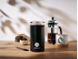 Black electric coffee grinder, coffee beans, ground coffee, and a French press on a wooden table.