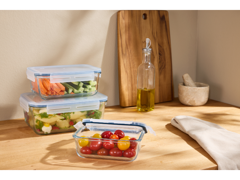 Glass food storage containers with vegetables and pasta on a wooden countertop.