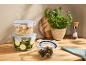 Glass containers with lids, filled with couscous, zucchini, and olives, on a wooden counter.