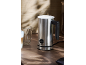 Silver milk frother on a wooden table, with a latte and an empty glass next to it.