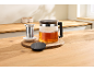 Glass teapot with tea, a tea infuser, and a lid on a wooden board in a kitchen.