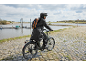 Cyclist in a rain poncho and backpack, riding on a cobblestone path by a river.