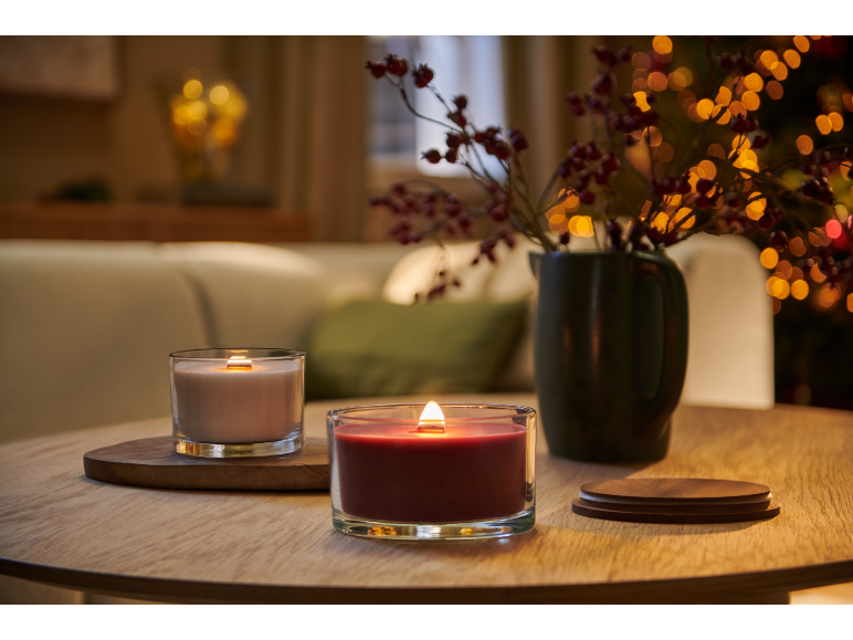 Two lit candles on a wooden table, with a vase of berries and blurred lights in the background.