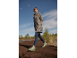 Woman in a leopard print jacket, blue jeans, and green boots walks on a muddy path.