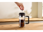 A hand presses down the plunger of a coffee press filled with coffee on a wooden counter.