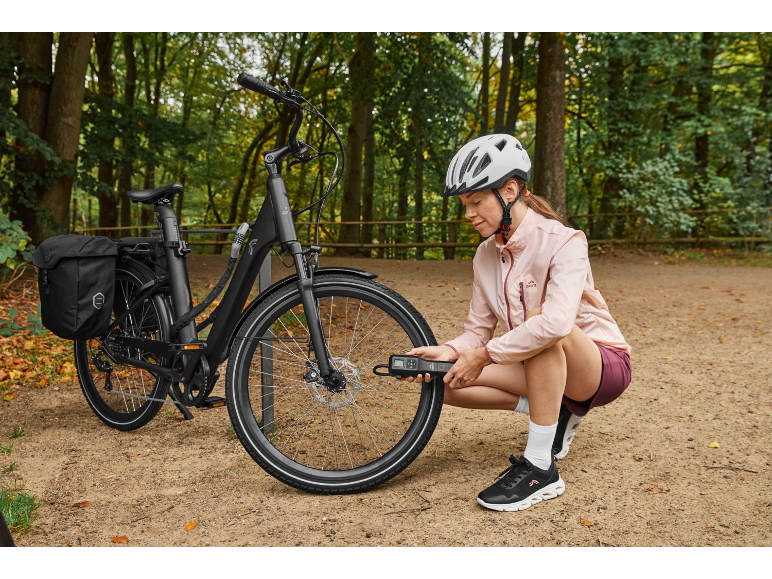Woman in helmet and pink jacket inflating tire of black e-bike with panniers in a park.