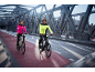 Two people in reflective cycling jackets and helmets riding electric bikes on a bridge.