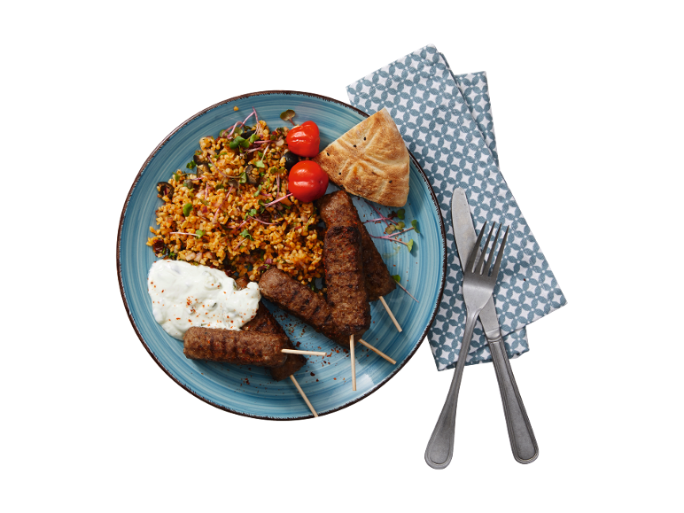 Kebab plate with bulgur, tomatoes, bread, and yogurt sauce, with cutlery and a napkin.