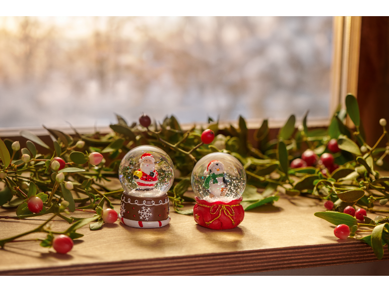 Two Christmas snow globes with Santa and a snowman, surrounded by mistletoe, on a windowsill.
