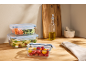 Glass containers with blue lids filled with vegetables and pasta on a wooden countertop.