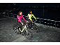 Two people in reflective cycling jackets riding electric bikes at night by a river.