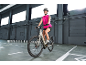 Smiling woman in pink sportswear and helmet, riding a grey electric bicycle in a warehouse.