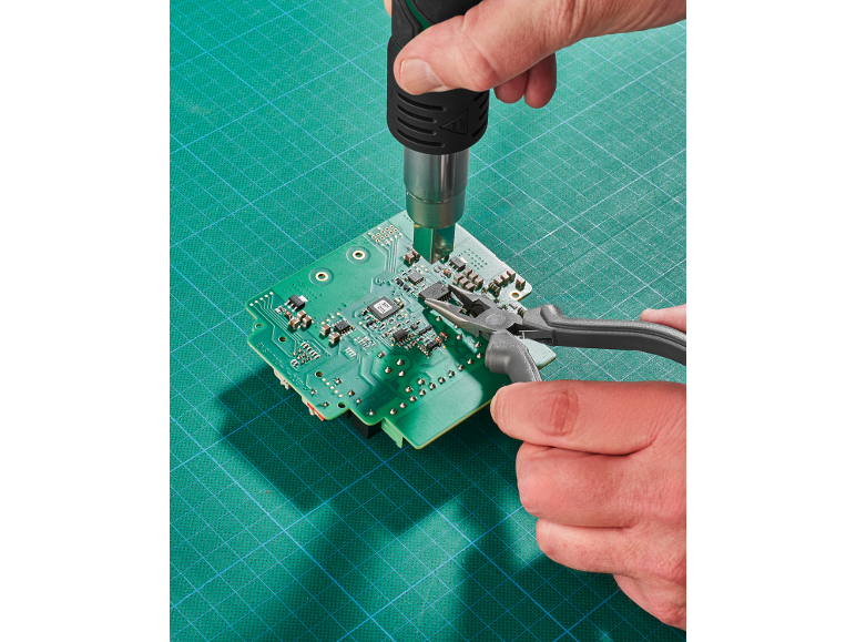 Hands using a heat gun and pliers to repair a green circuit board.