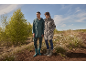 Man and woman in rain jackets and boots stand in a field with white flowers.