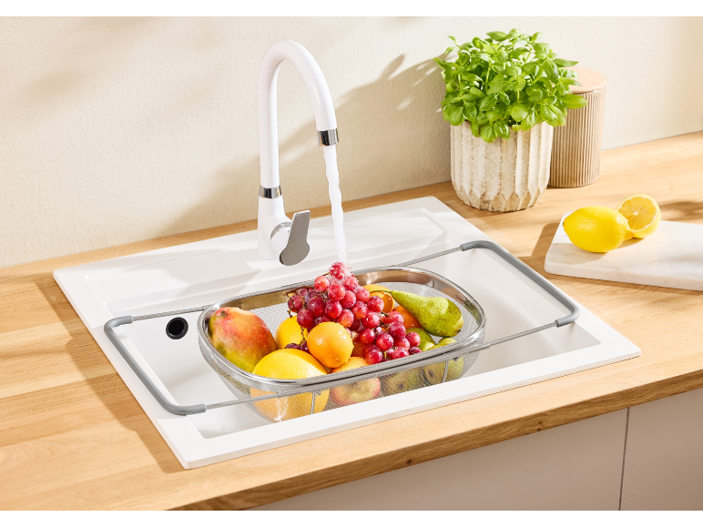 White kitchen faucet with fruit in a colander over a sink and a plant.