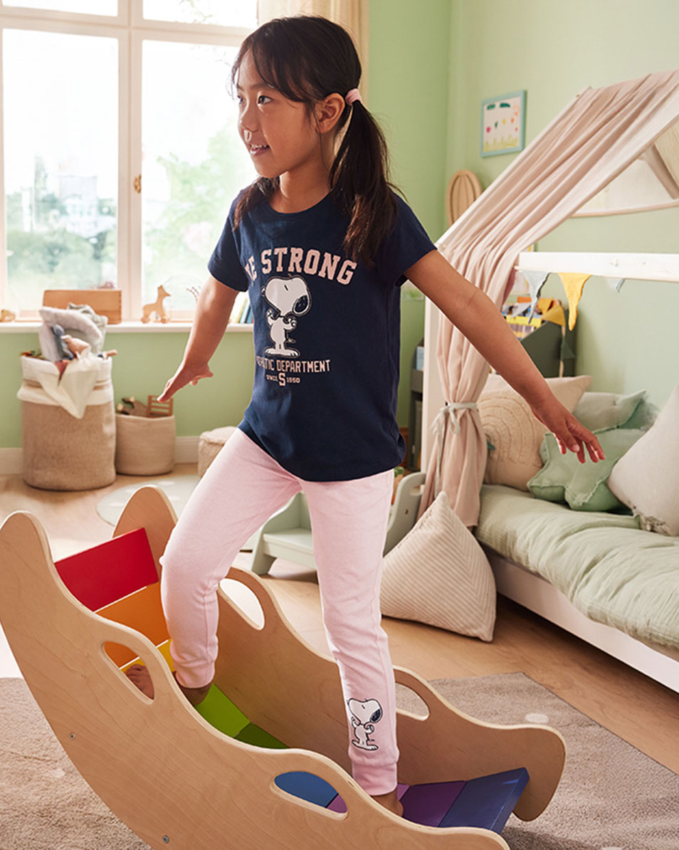 Girl in a navy t-shirt with a cartoon dog print and pink leggings playing on a wooden rocker.