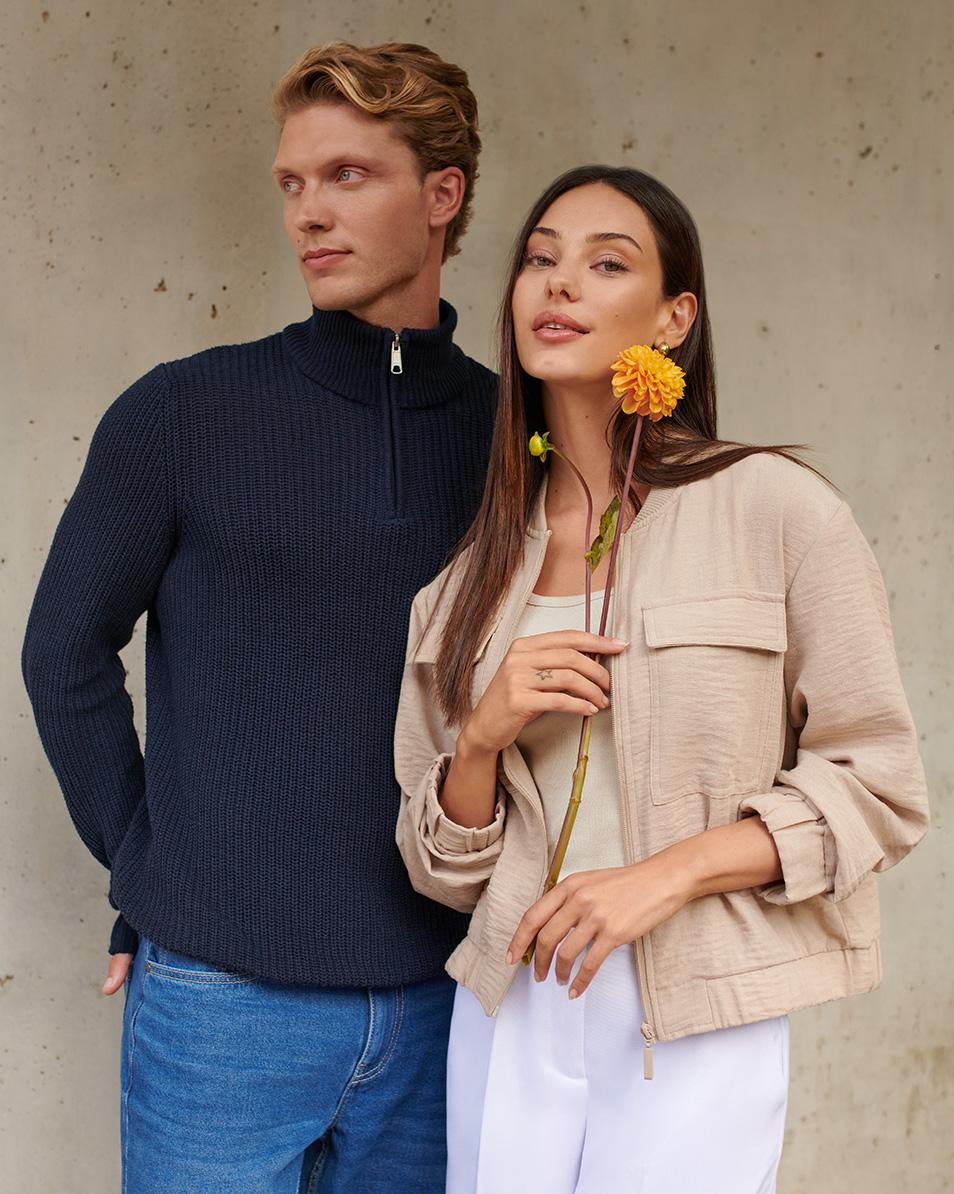 Man in dark blue sweater and jeans, woman in beige jacket and white pants, holding a flower.