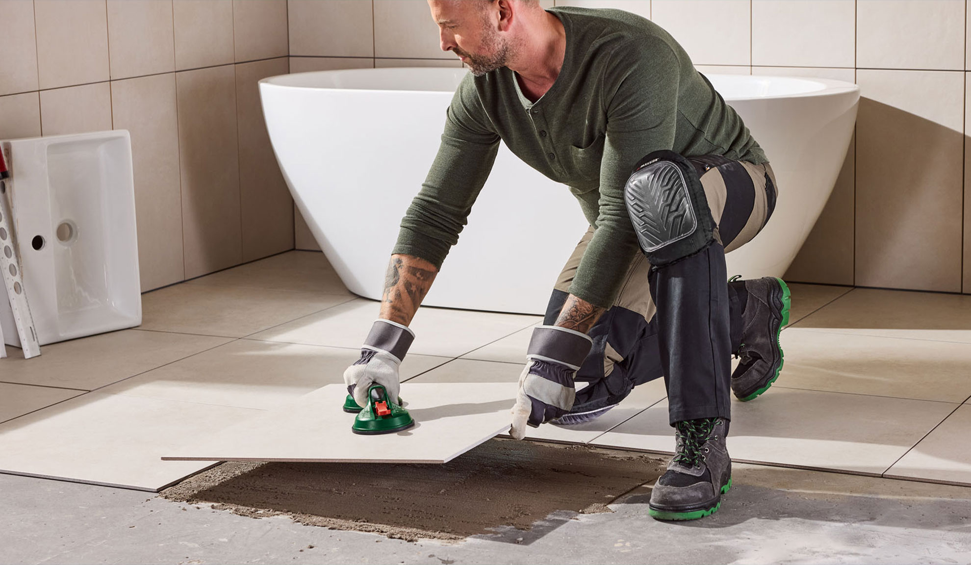 Man laying tiles in a bathroom using a tile suction cup and knee pads.