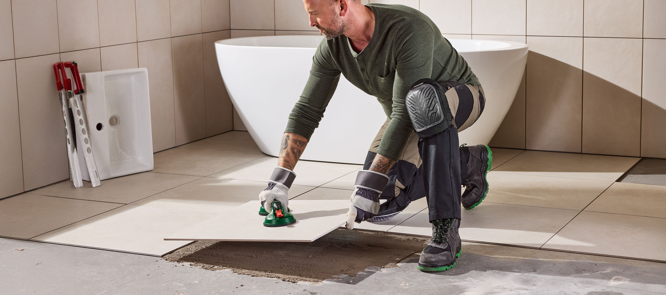 Man tiling a bathroom floor with large light-colored tiles, wearing knee pads and workwear.