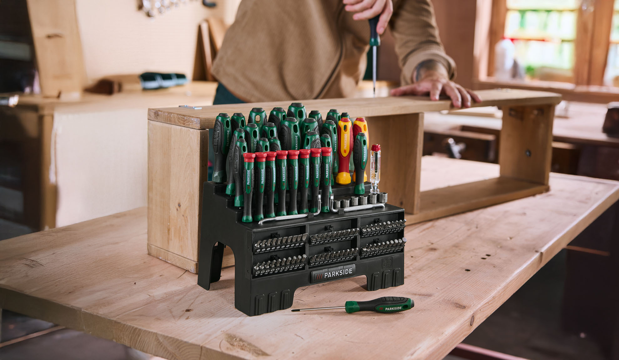 Screwdriver and bit set on a wooden table, person assembling a wooden shelf.