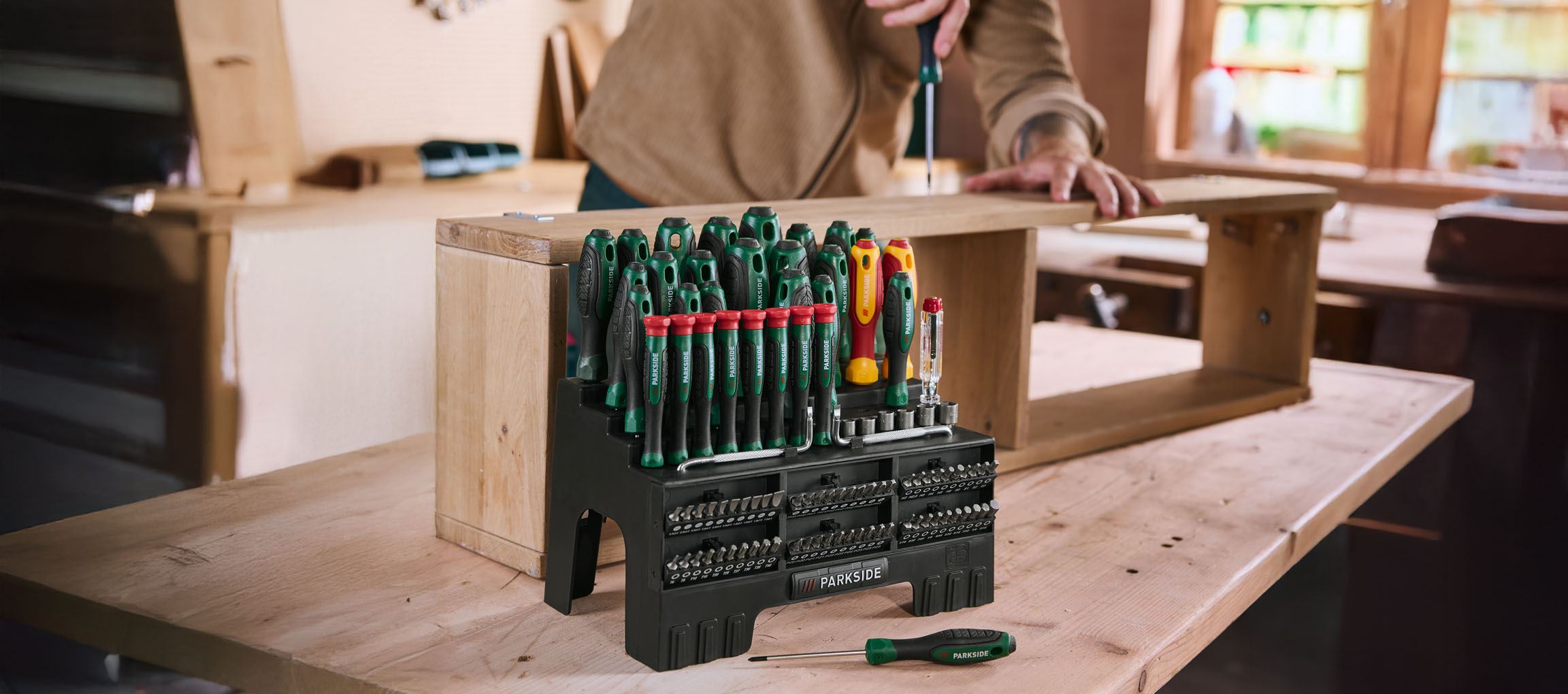 A large screwdriver and bit set on a wooden workbench, with a person assembling furniture.