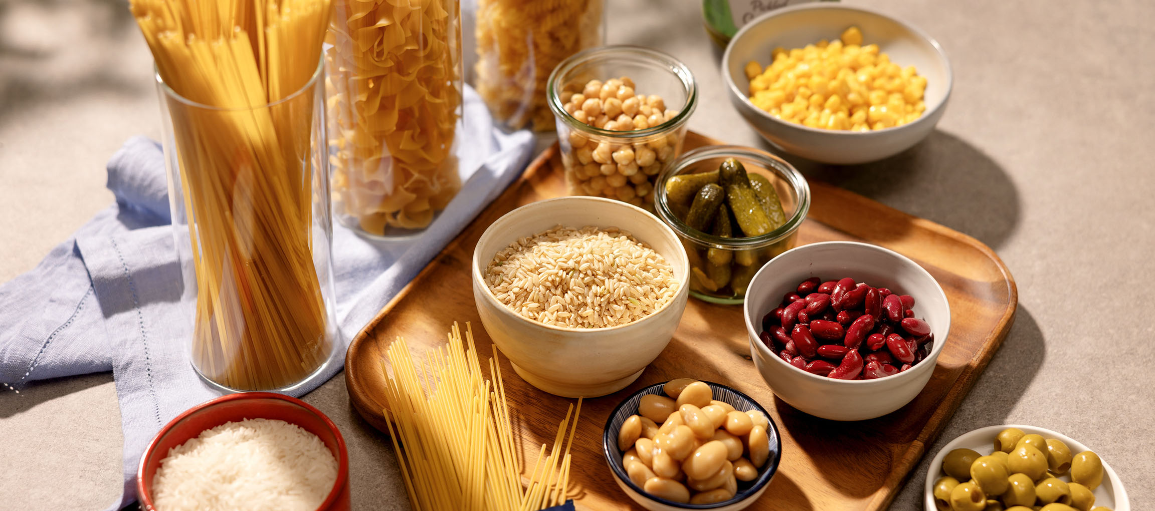 Various pantry staples including pasta, rice, beans, chickpeas, corn, pickles, and olives.