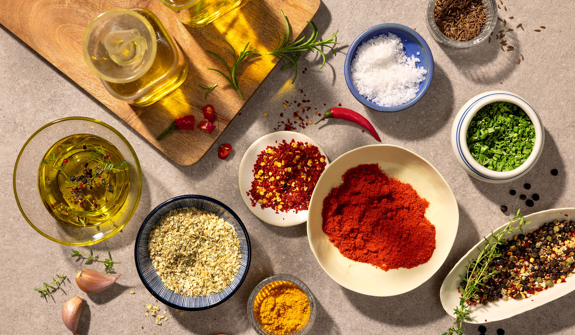 Overhead shot of various spices, herbs, and olive oil on a grey surface.