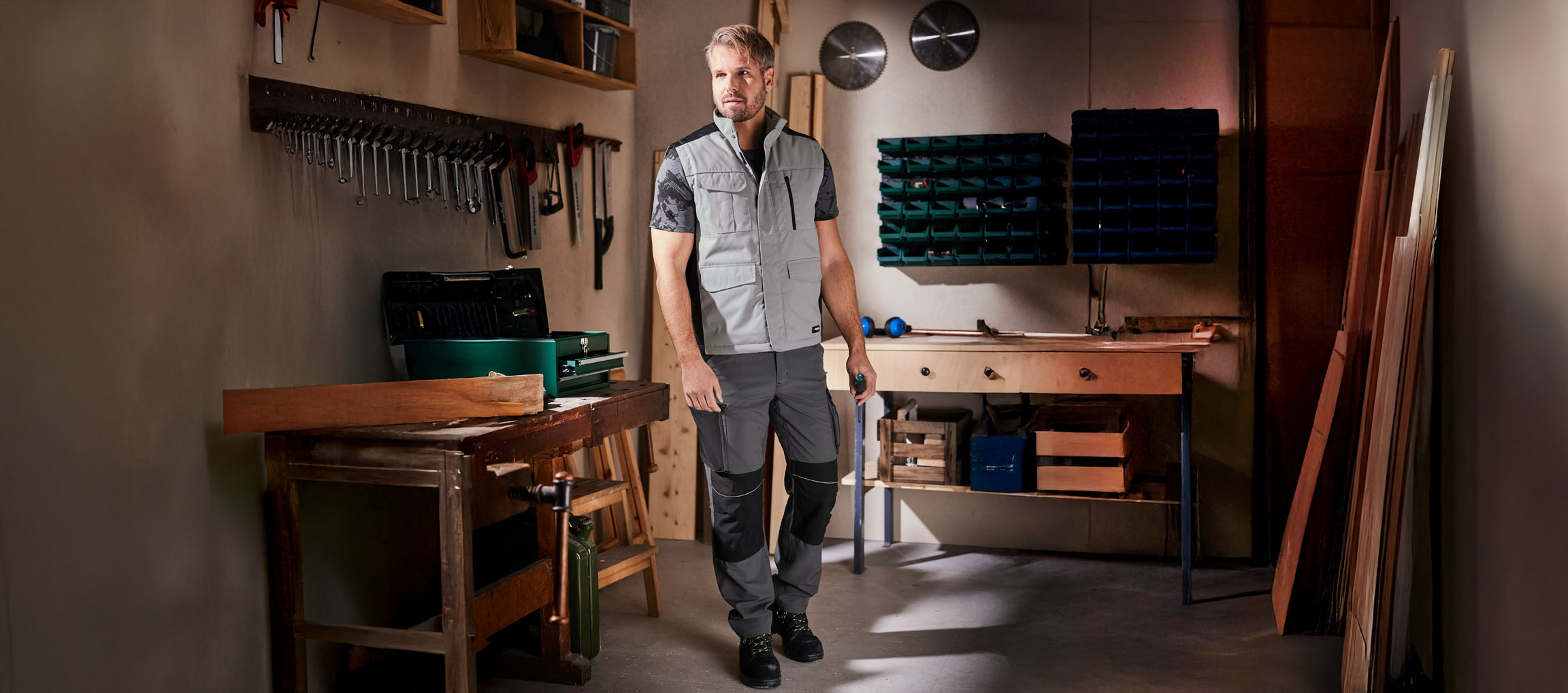 A man in workwear and vest stands in a workshop.