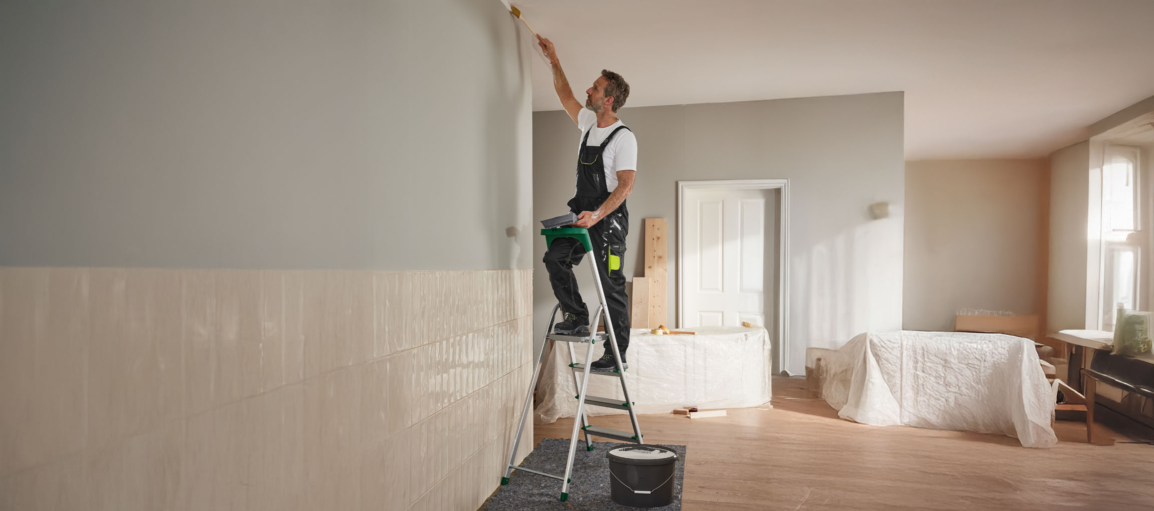 A man paints a wall with a brush, standing on a stepladder.