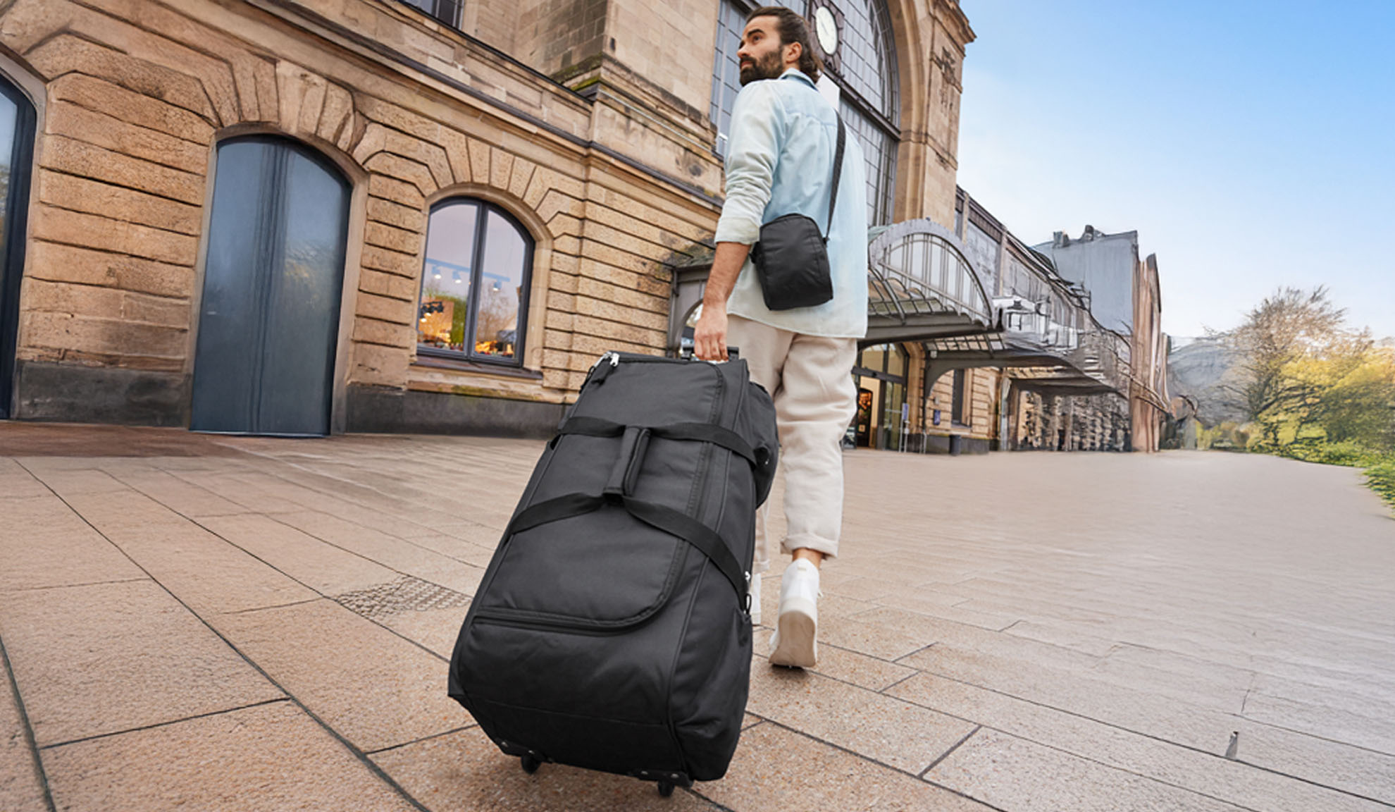 A man walks with a wheeled duffel bag near a train station.