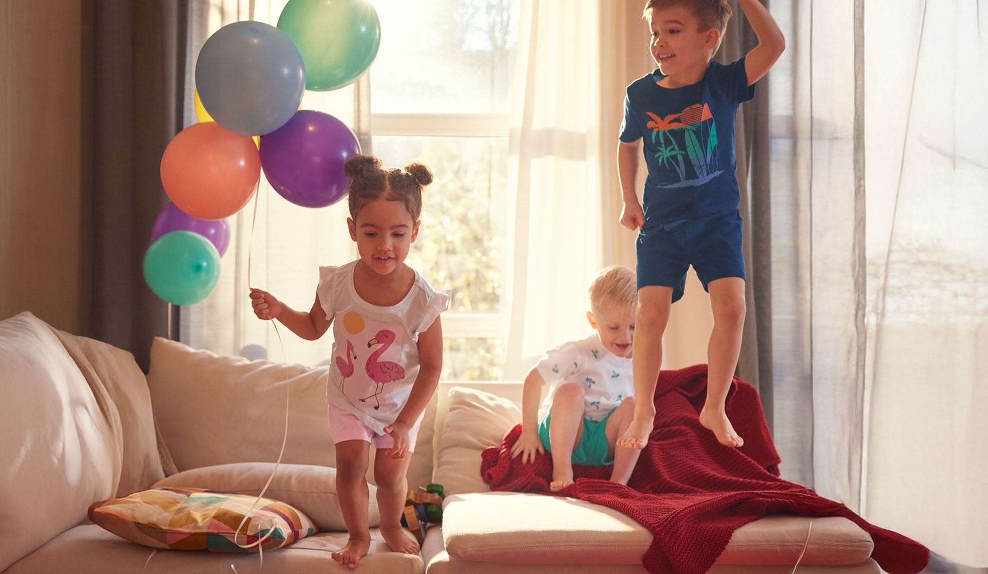 Three children joyfully jump on a sofa with colorful balloons.