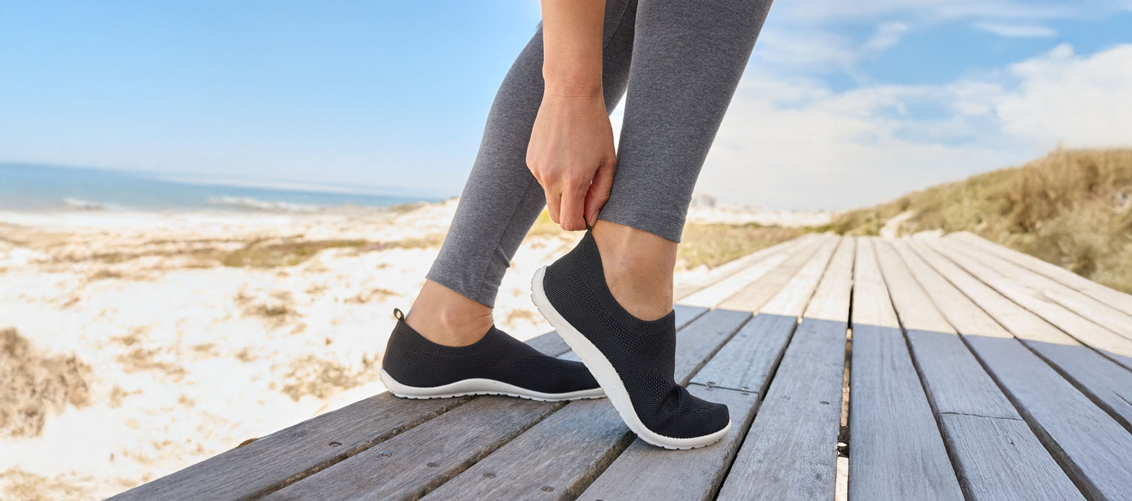 Woman putting on black slip-on sneakers by the beach.
