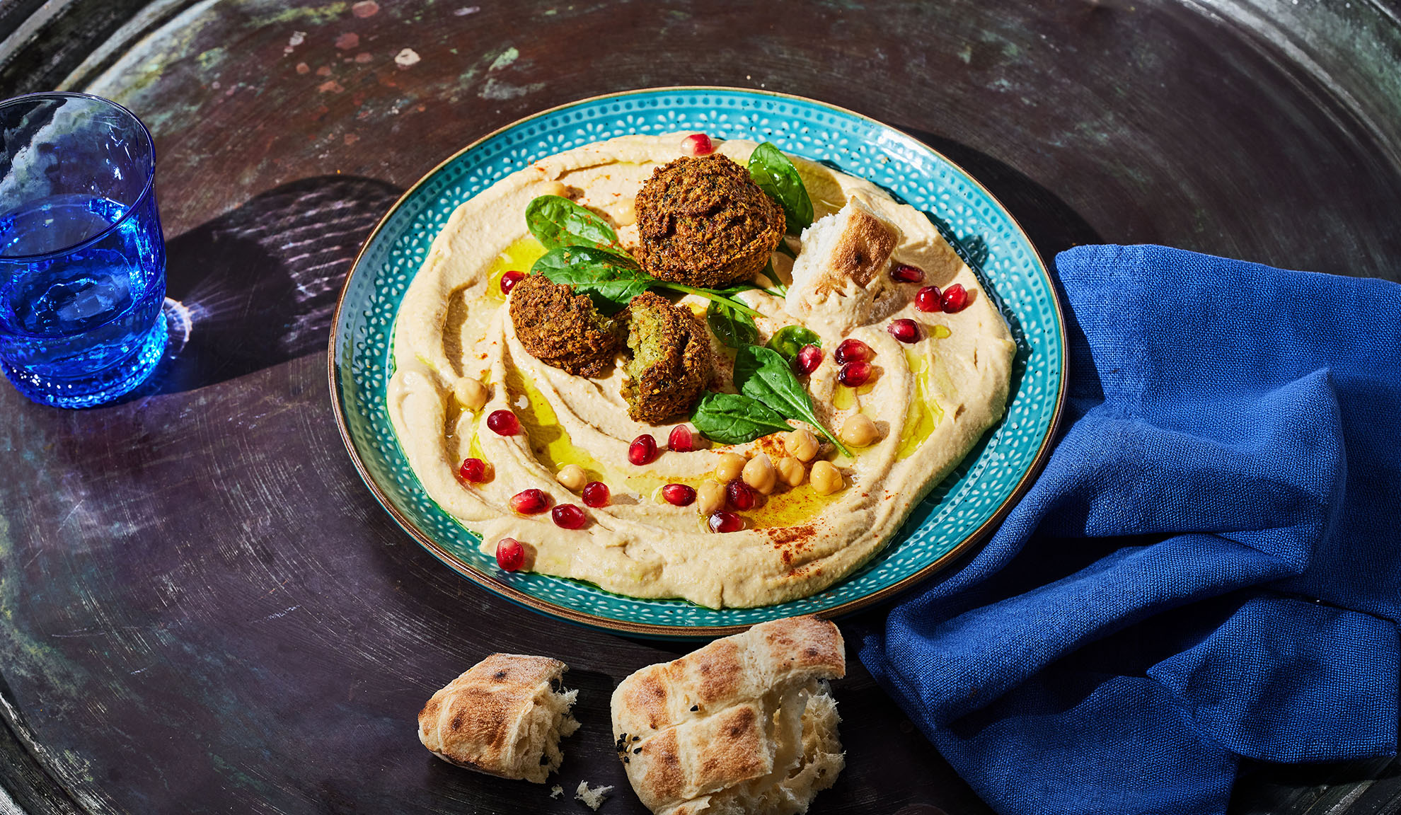 Hummus with falafel, pomegranate seeds, and pita bread on a dark table.