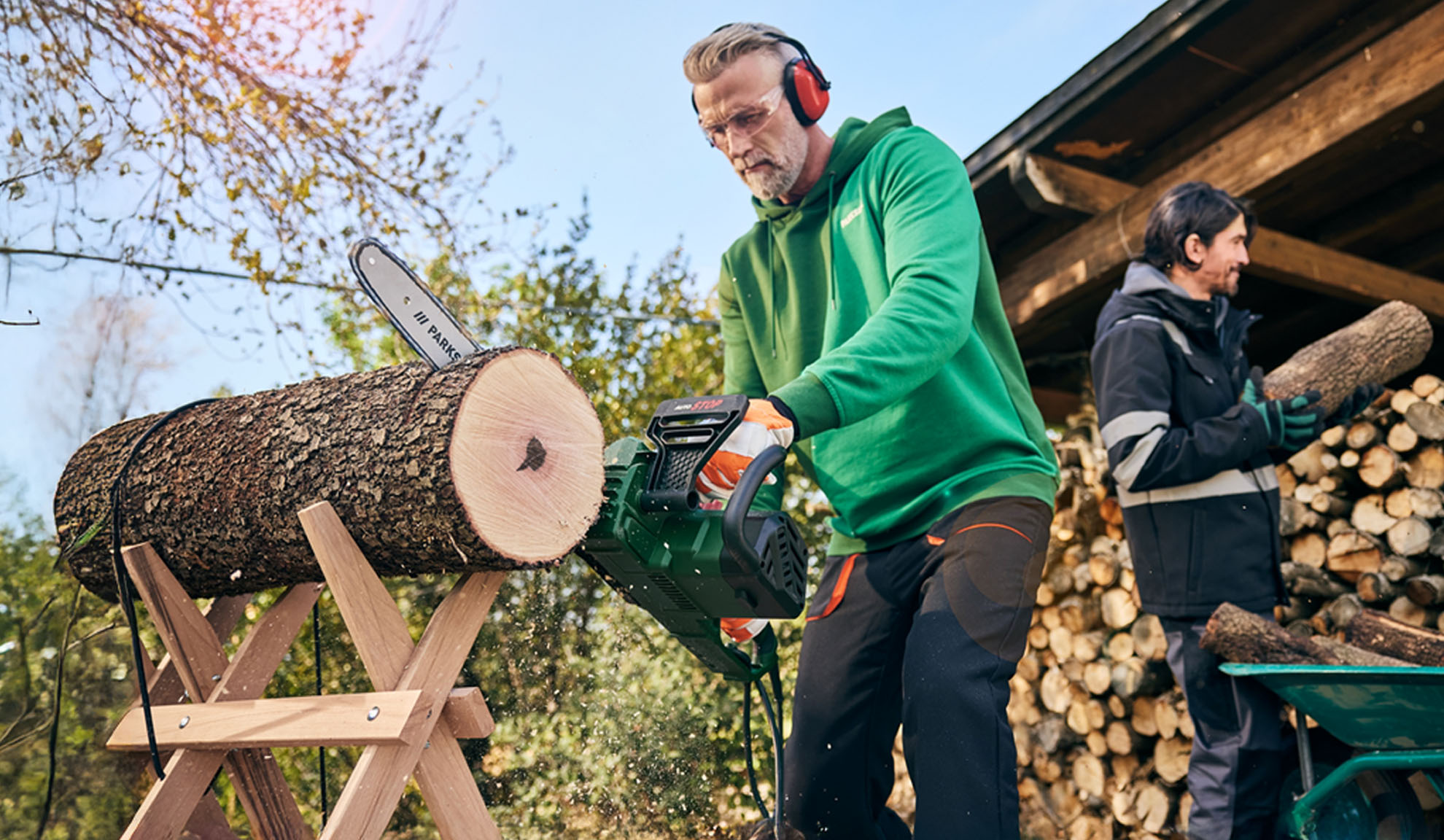 Man cutting log with a chainsaw while another man stacks firewood.
