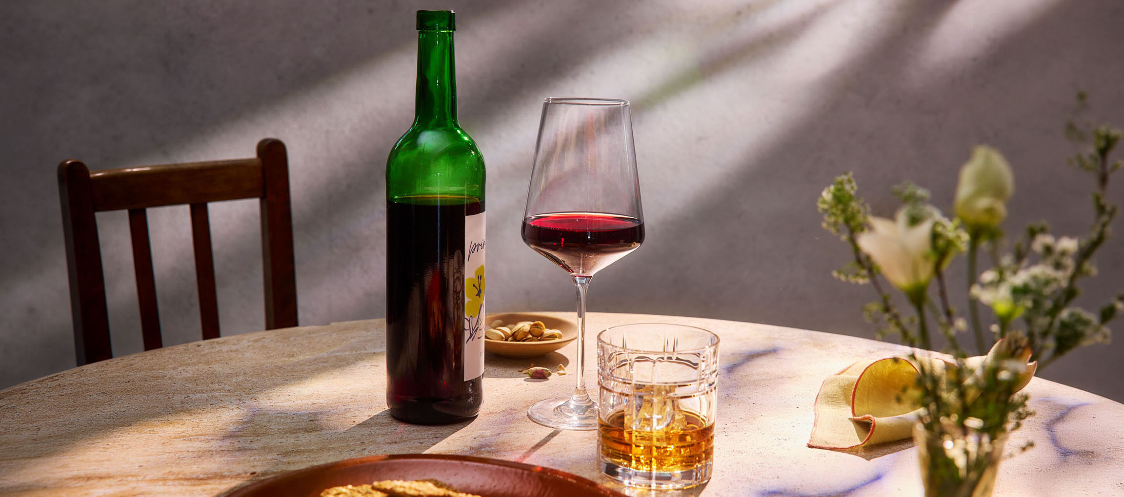 Bottle of red wine, glass of wine, and whiskey on a table, with a chair and flowers.