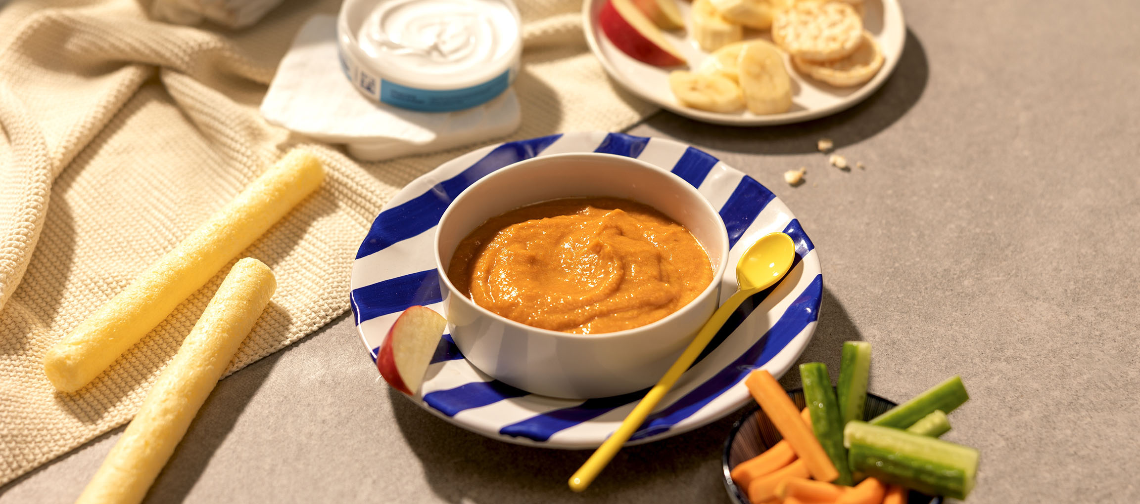 Baby food puree with snacks like fruit, crackers, and vegetable sticks.