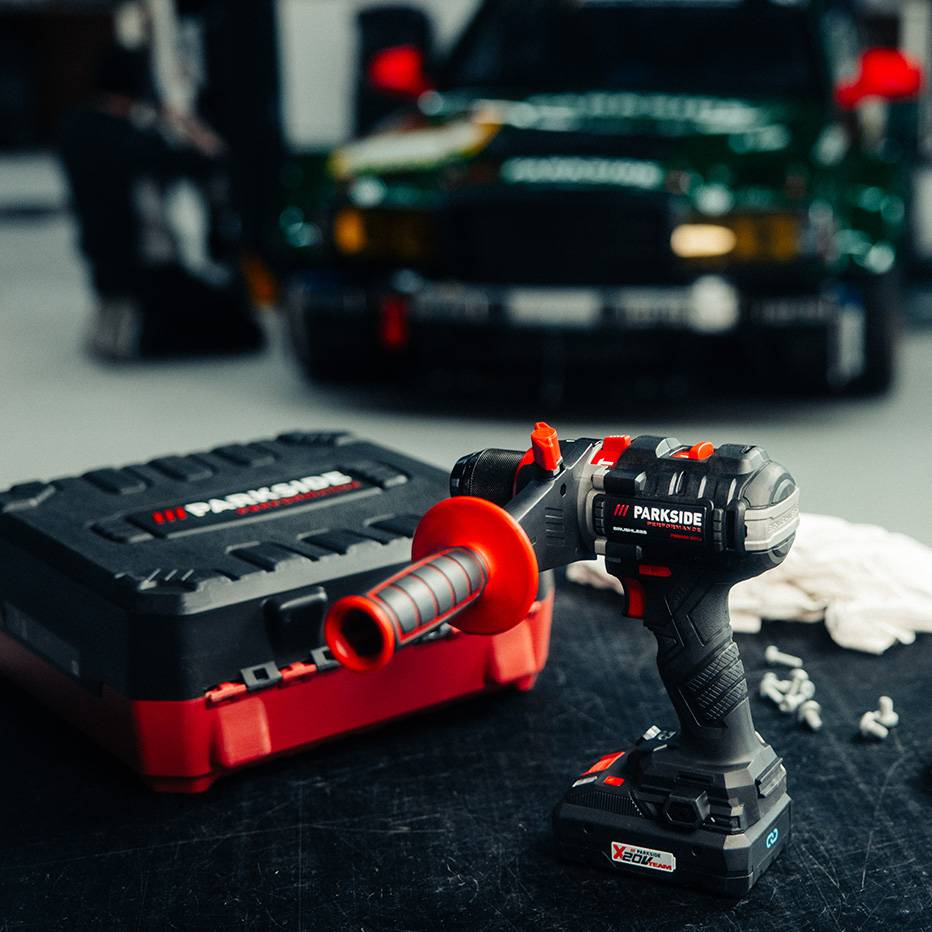 Cordless drill with case and accessories on a workbench, with a car in the background.