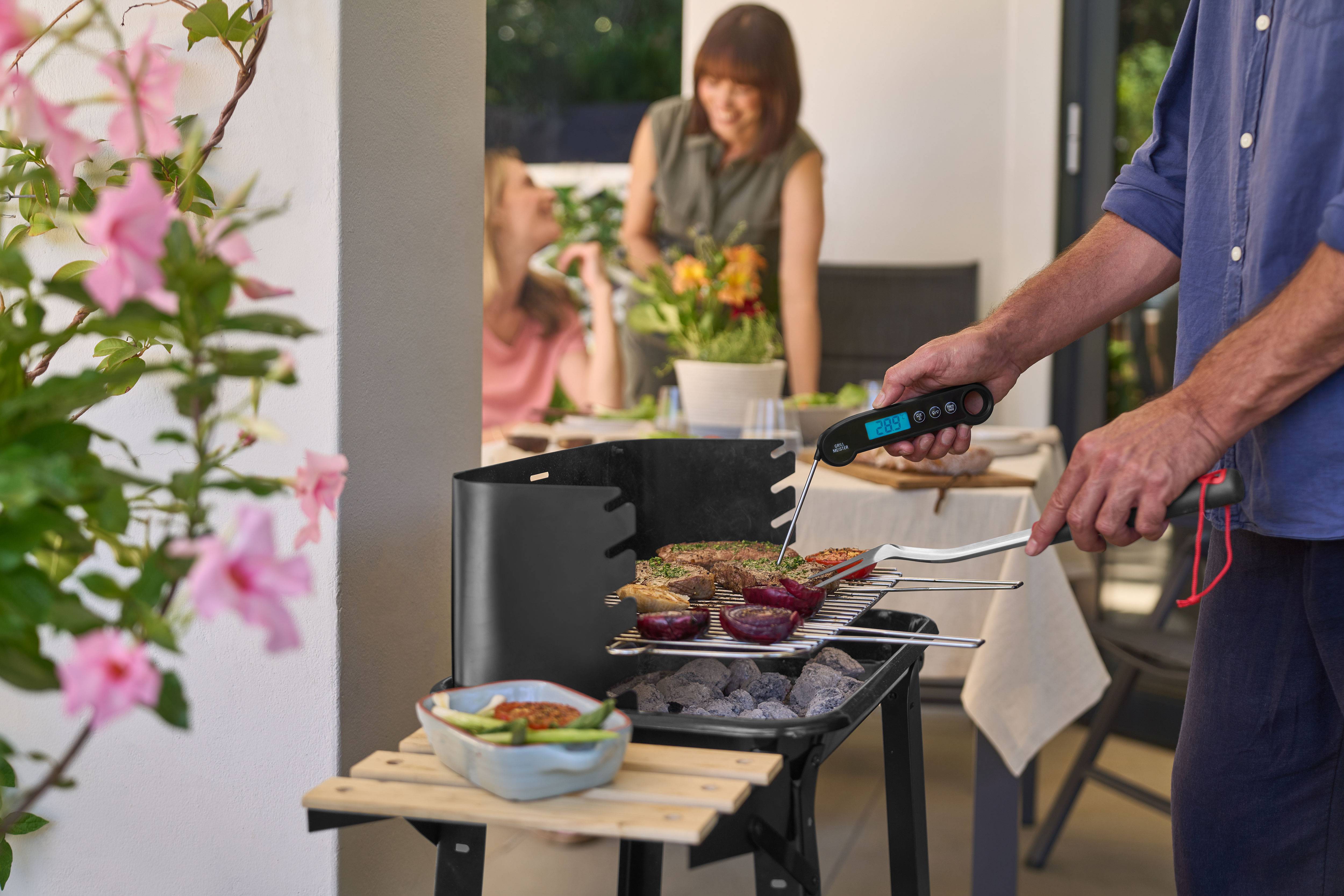 Man grilling meat and vegetables using a meat thermometer, with women in the background.