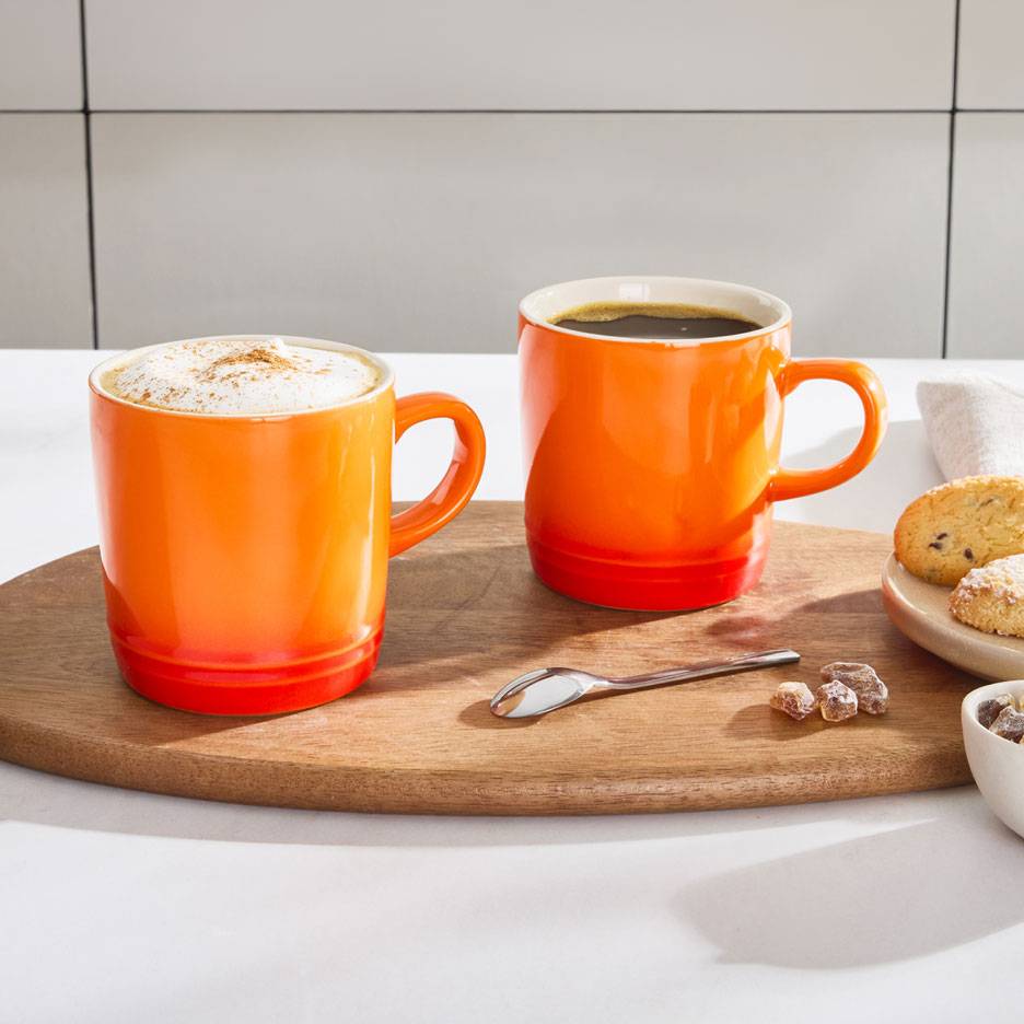 Two orange coffee mugs on a wooden tray, one with frothy coffee, the other with black coffee.
